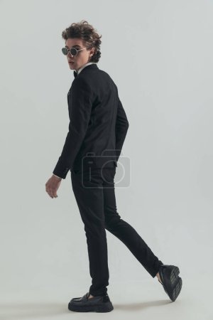 Téléchargez les photos : Rear view picture of young groom in elegant suit looking behind and walking in front of grey background - en image libre de droit