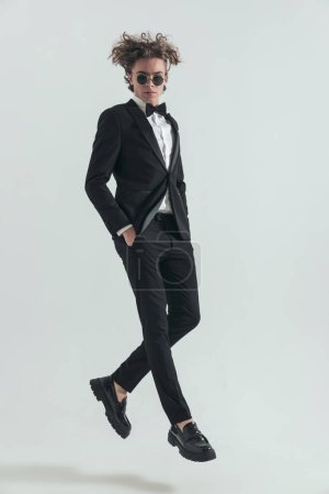 Foto de Full body picture of curly hair man in tuxedo jumping in the air with hands in pockets in front of grey background in studio - Imagen libre de derechos