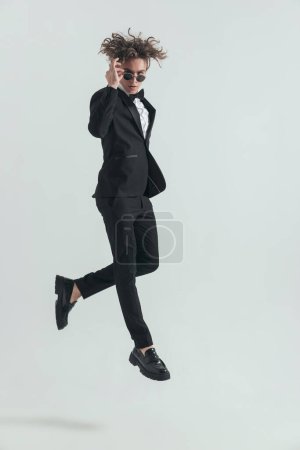 Photo for Fashion elegant groom in tuxedo adjusting sunglasses while jumping in front of grey background in studio - Royalty Free Image