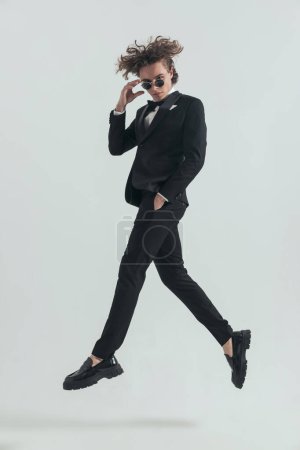 Photo for Caucasian man with blonde curly hair wearing black tuxedo leaping up in the air with hand in pocket and adjusting glasses on grey background - Royalty Free Image