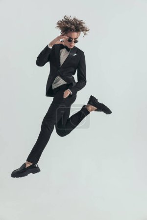 Foto de Full body picture of curly hair man in tuxedo jumping in the air with one hand in pocket and adjusting sunglasses with the other one on grey background - Imagen libre de derechos