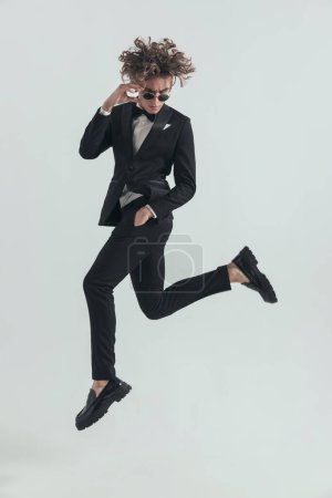 Photo for Side view of sexy elegant man jumping in the air with hand in pockets and adjsuting sunglasses in front of grey background in studio - Royalty Free Image