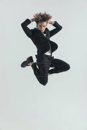 Photo for Fashion young man with curly hair jumping in the air with hands behind head in front of grey background in studio - Royalty Free Image