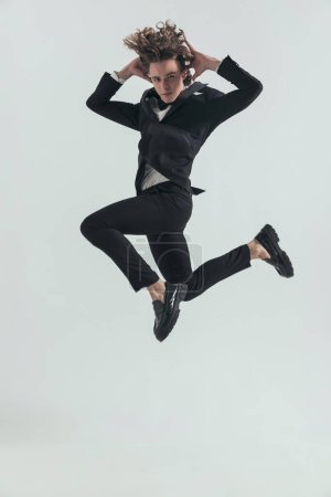 Téléchargez les photos : Full picture of elegant man in tuxedo jumping in the air with hands behind bead and posing in a cool way in front of grey background - en image libre de droit