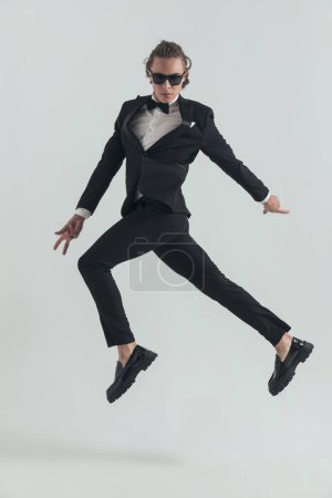 Photo for Elegant man in tuxedo jumping in the air in a dynamic way and posing in a fashion manner in front of grey background in studio - Royalty Free Image