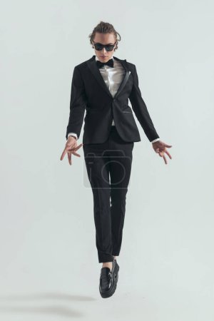Photo for Young groom in tuxedo with sunglasses leaping up in the air and making hand gestures on grey background - Royalty Free Image