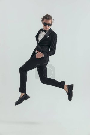 Photo for Side view of sexy man in elegant tuxedo with sunglasses jumping in the air and making thumbs up gesture on grey background - Royalty Free Image