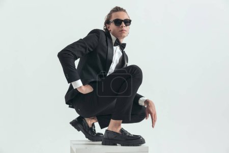 Photo for Side view picture of handsome young elegant man crouching with hand in pocket and posing on grey background - Royalty Free Image