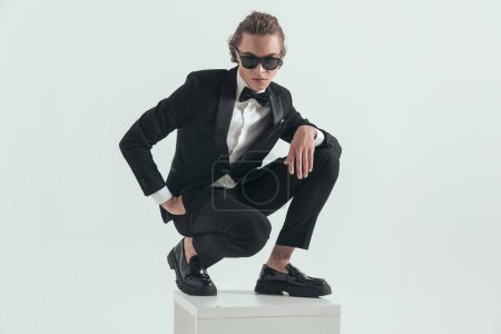 Photo for Sexy elegant man with blone hair crouching with hand in pockets while posing with the other hand above knee in front of grey background - Royalty Free Image