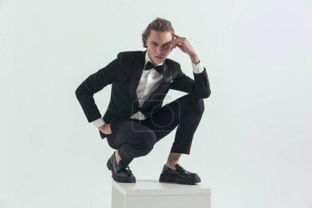 Photo for Man with blue eyes wearing black tuxedo, crouching with elbow on knee, touching temple and holding hand in pocket in front of grey background - Royalty Free Image