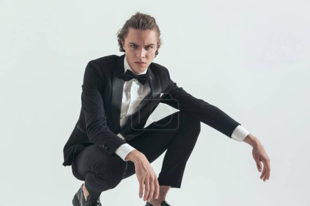 Photo for Picture of sexy elegant best man in black tuxedo crouching with elbows on knees and posing in a confident and serious manner on grey background - Royalty Free Image