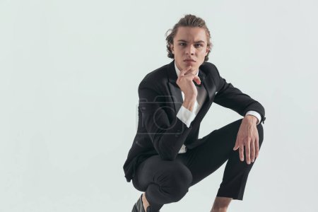Téléchargez les photos : Pensive young groom in black tuxedo touching chin and thinking while crouching with elbow on knee in front of grey background in studio - en image libre de droit