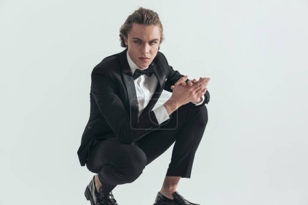 Téléchargez les photos : Handsome man in elegant black tuxedo with blue eyes crouching and rubbing palms in front of grey background in studio - en image libre de droit