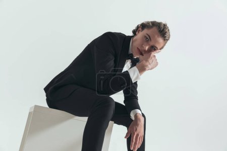 Photo for Attractive young man with blue eyes wearing tuxedo and posing with arms in fashion pose while sitting on grey background - Royalty Free Image