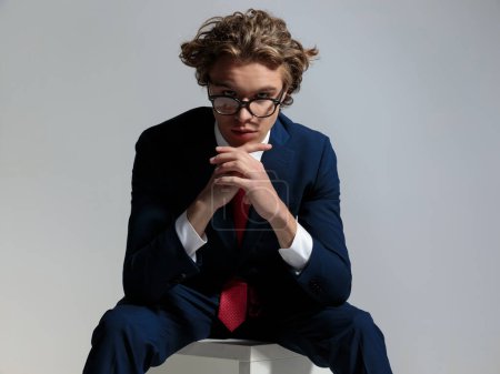 Téléchargez les photos : Portrait of sexy seated businessman with long curly hair holding elbows on knees and posing with hands together in front of grey background in studio - en image libre de droit