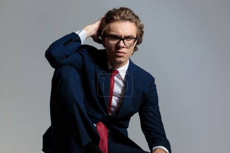 Téléchargez les photos : Cool businesssman with glasses adjusting long curly hair while posing with knee up and elbow above on grey background in studio - en image libre de droit