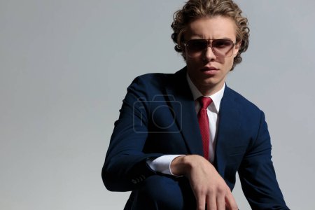 Photo for Portrait of sexy businessman in suit with sunglasses posing with arm on knee in front of grey background in studio - Royalty Free Image