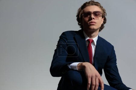Téléchargez les photos : Portrait of young man with curly hair wearing elegant suit and sunglasses and posing with arm over knee in front of grey background in studio - en image libre de droit