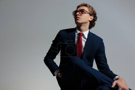 Foto de Handsome man with curly hair looking up side and posing with hand in pocket while holding leg over knee on grey background in studio - Imagen libre de derechos