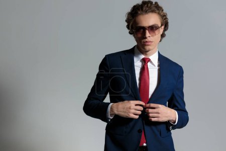 Foto de Young man with curly long hair with sunglasses adjusting and buttoning suit in front of grey background in studio - Imagen libre de derechos