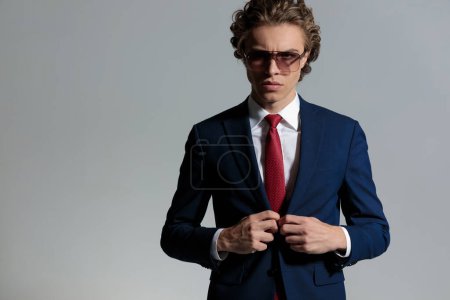 Téléchargez les photos : Portrait of confident young businessman wearing sunglasses and unbuttoning suit in front of grey background in studio - en image libre de droit