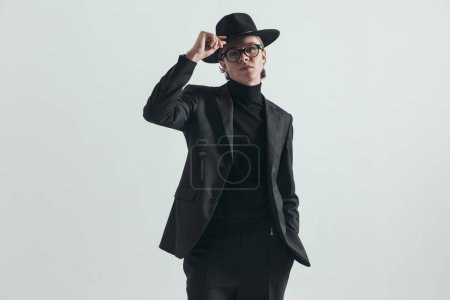 Téléchargez les photos : Attractive man with cool outfit posing with hand in pocket while adjusting hat in front of grey background in studio - en image libre de droit