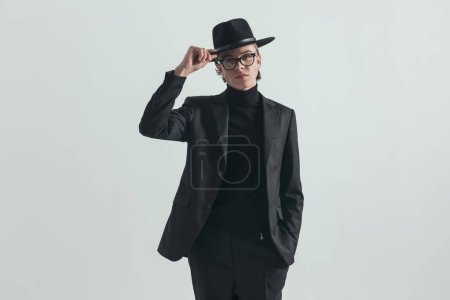 Foto de Portrait of cool fashion man with glasses adjusting hat and posing with hand in pockets in front of grey background in studio - Imagen libre de derechos