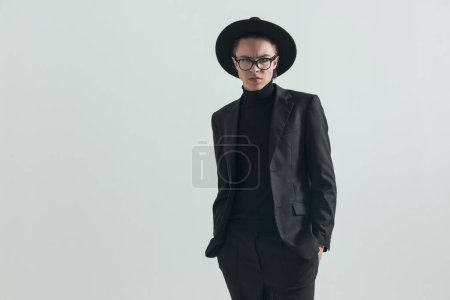 Téléchargez les photos : Arrogant young man wearing full black outfit with hat and glasses holding hands in pockets in front of grey background in studio - en image libre de droit