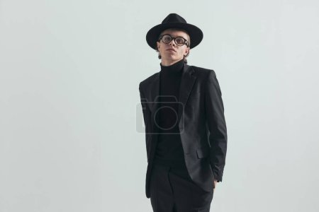 Foto de Portrait of cool man with turtleneck and black suit posing with hands in pockets, wearing black hat and glasses, being confident on grey background - Imagen libre de derechos