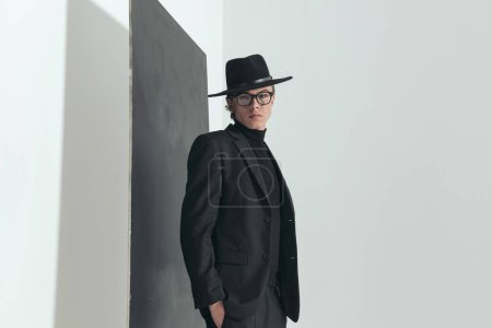 Foto de Side view picture of elegant man with black suit posing with hands in pockets in front of grey background in studio - Imagen libre de derechos