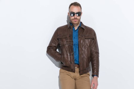 Foto de Sexy casual man with brown leather jacket and sunglasses posing with hand in pocket in front of white background in studio - Imagen libre de derechos
