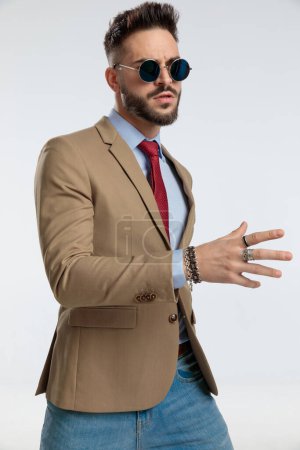 Photo for Portrait of a sexy businessman with tough attitude looking away, standing, wearing sunglasses against gray studio background - Royalty Free Image