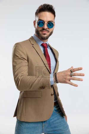 Photo for Portrait of a attractive businessman posing with one open hand, standing, wearing sunglasses against gray studio background - Royalty Free Image