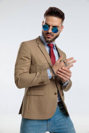 Foto de Portrait of a young businessman holding his hand and looking away with tough attitude, standing, wearing sunglasses against gray studio background - Imagen libre de derechos