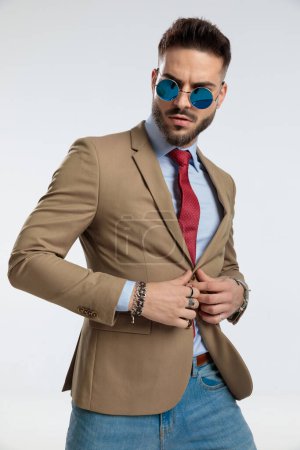 Foto de Portrait of a handsome businessman holding hands on his jacket's button, standing, wearing sunglasses against gray studio background - Imagen libre de derechos