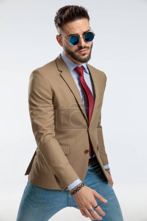 Foto de Portrait of a attractive businessman holding one hand in front and behind with tough attitude, sitting, wearing sunglasses against gray studio background - Imagen libre de derechos