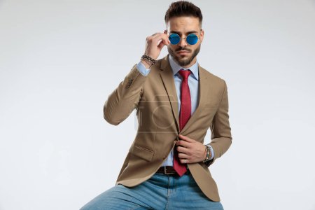 Photo for Portrait of a sexy businessman fixing his jacket, sitting, arranging sunglasses against gray studio background - Royalty Free Image