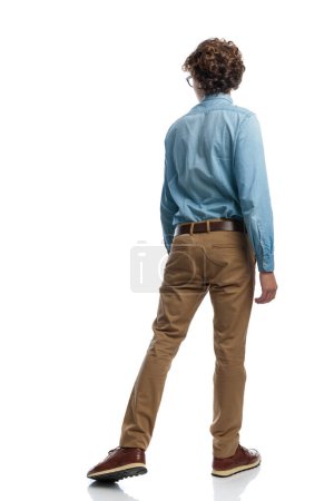 Foto de Imagen de cuerpo completo de un hombre casual sexy caminando de una manera y mirando a la otra, de pie, usando anteojos contra el fondo blanco del estudio - Imagen libre de derechos