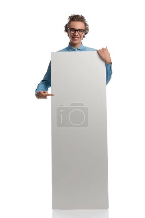 Téléchargez les photos : Full body picture of sexy casual man presenting billboard while pointing his finger, standing, wearing eyeglasses against white studio background - en image libre de droit