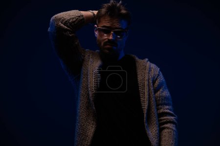 Téléchargez les photos : Unshaved cool man with cardigan and sunglasses holding hand behind neck and posing on dark background - en image libre de droit