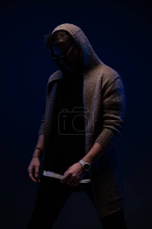 Photo for Stylish young guy with sunglasses wearing hoodie and looking down while posing in a fashion way on dark background - Royalty Free Image