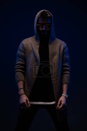 Téléchargez les photos : Dramatic cool guy with sunglasses wearing wool cardigan and posing on dark background - en image libre de droit