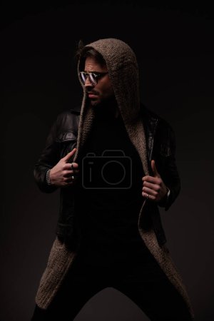 Téléchargez les photos : Sexy young man with wool cardigan and glasses arranging leather jacket and looking to side while confidently posing on grey background - en image libre de droit