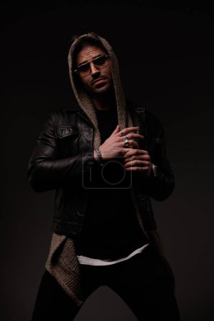 Photo for Confident cool man with hoodie wearing leather jacket and wool cardigan and pulling fingers on grey background - Royalty Free Image