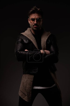 Téléchargez les photos : Confident man with cool hairstyle crossing arms and posing while wearing leather jacket and wool cardigan on grey background - en image libre de droit