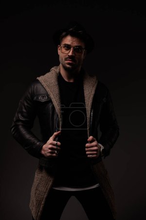 Téléchargez les photos : Sexy barbu gars avec des lunettes tirant et ajustant veste en cuir et cardigan de laine tout en regardant loin sur fond gris - en image libre de droit