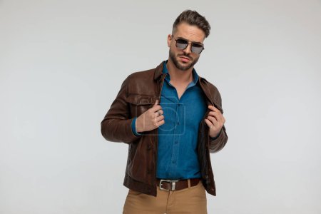 Photo for Portrait of a handsome casual man adjusting his jacket with tough style, standing, wearing sunglasses against gray studio background - Royalty Free Image