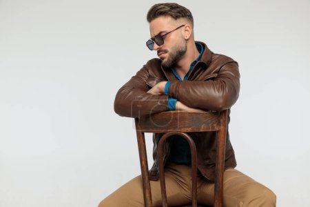 Photo for Portrait of a handsome casual man resting his arms and being dramatic, sitting on a chair, wearing sunglasses against gray studio background - Royalty Free Image