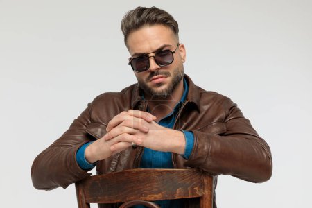 Foto de Portrait of a attractive casual man resting his arms together with tough attitude, sitting on a chair, wearing sunglasses against gray studio background - Imagen libre de derechos