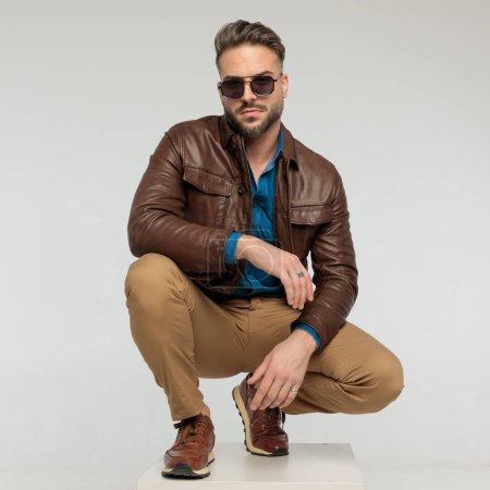 Photo for Portrait of a sexy casual man resting his arms and squatting, standing, wearing sunglasses against gray studio background - Royalty Free Image